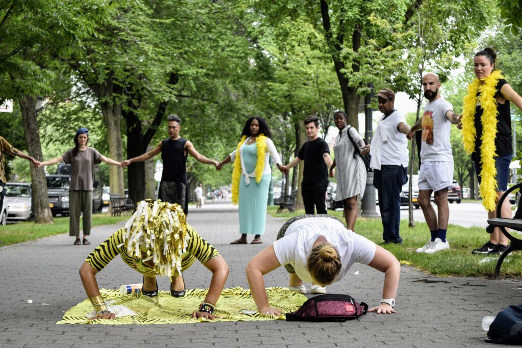 Two women in push up position outside. Ayana, the person on the left, is dressed in yellow tiger print with a pom pom on their head.