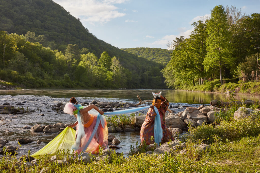 two figures dressed up in tule, one tugging the other. They're standing on the edge of a river during a perfect sunny day.