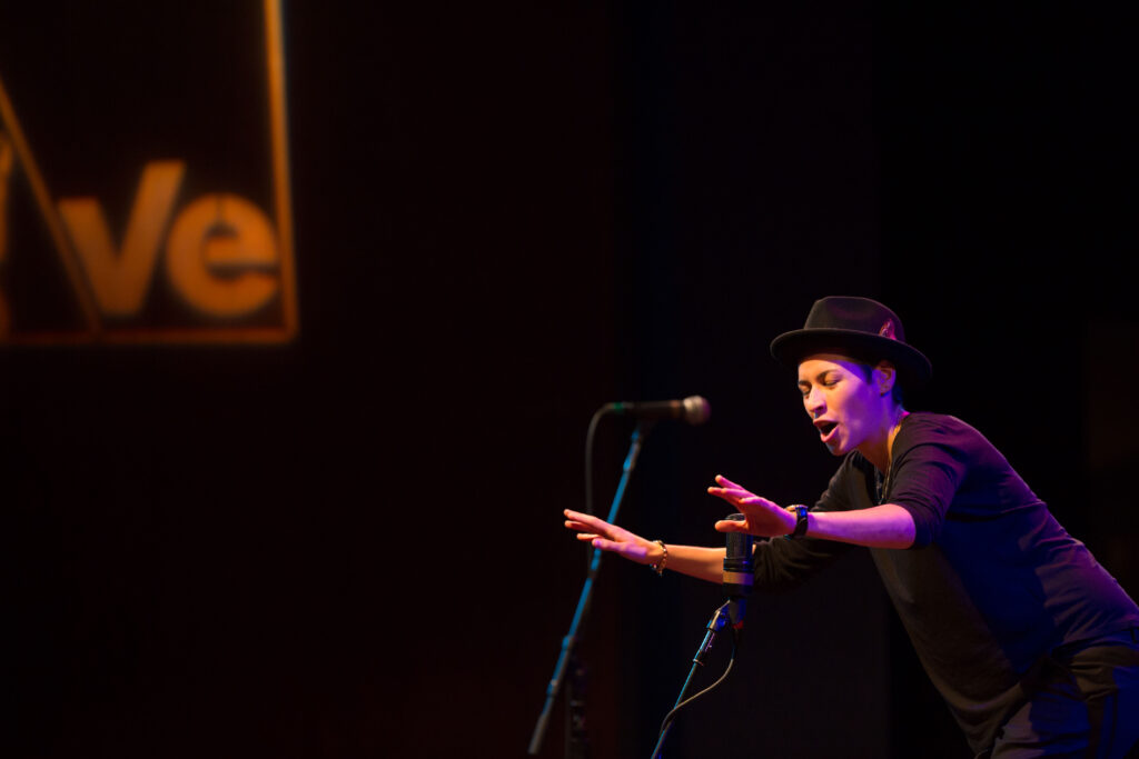 A person on a dark stage gesturing and speaking into a microphone