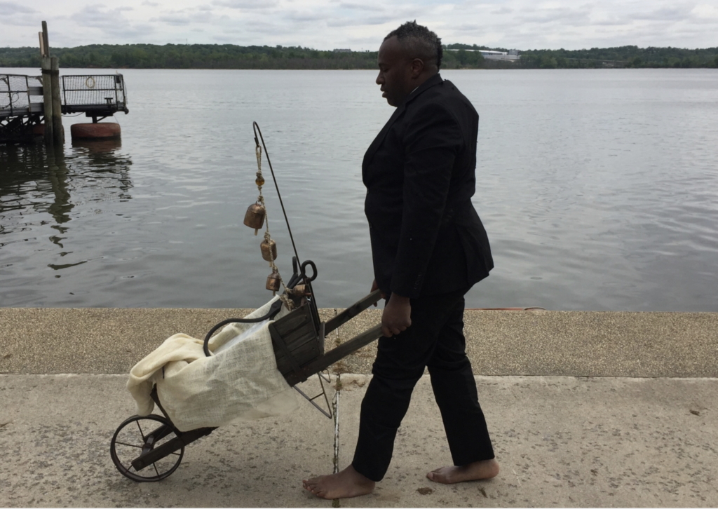 A Black man in a suit pushing a cart along a sidewalk beside a body of water.