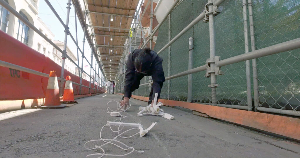 someone building with small white blocks and white thread on the ground under scaffolding