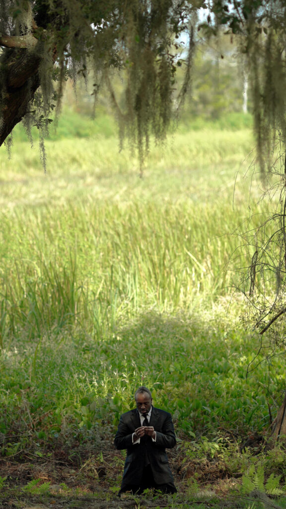 A photo of a Black man in a suit kneeling beneath some trees.