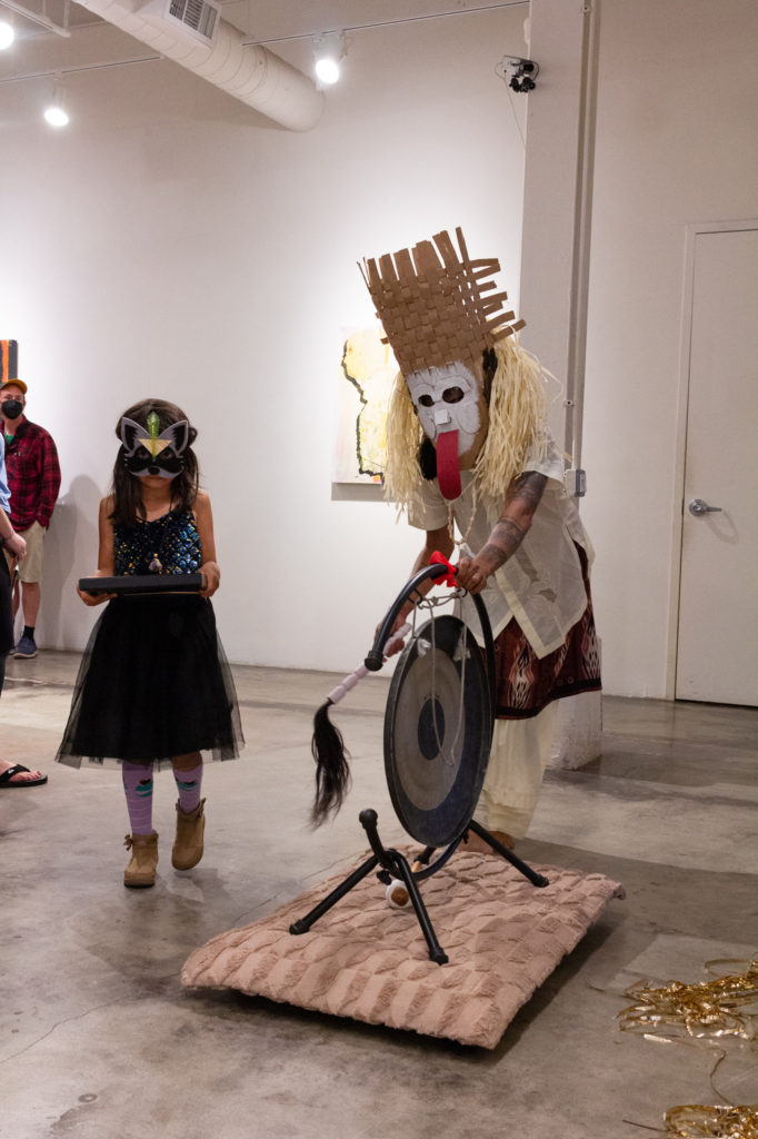 Person in a cardboard mask with white face and long red tongue pushing a gong, a child in a raccoon mask beside.