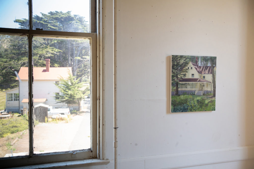 A painting of a house hung next to a window with a similar view