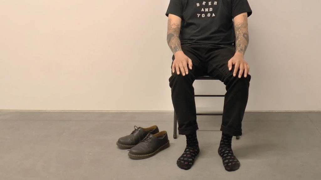 Photo of a person from the neck down sitting on a chair, their shoes on the floor beside them