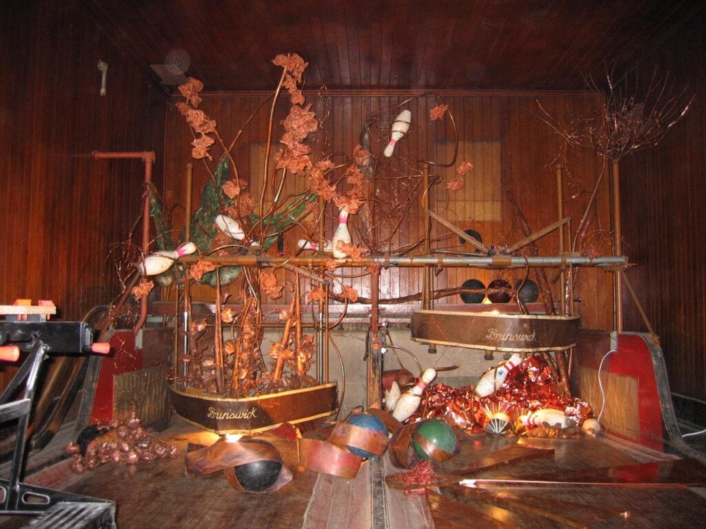 An art installation of bowling balls, bowling pins, and copper colored structure on a bowling lane.