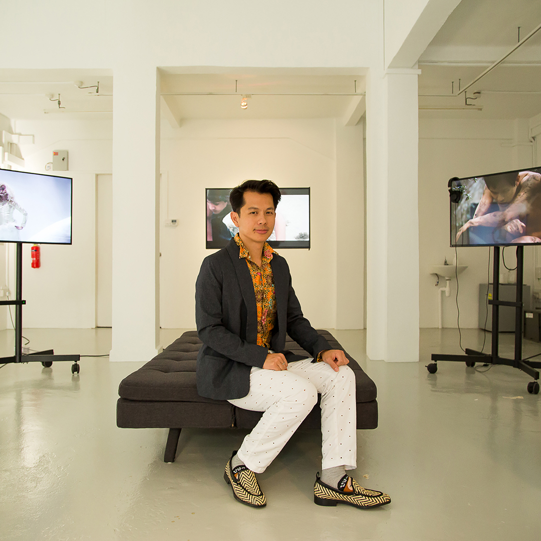 Photo of a person sitting on a bench in a white room, video monitors behind them
