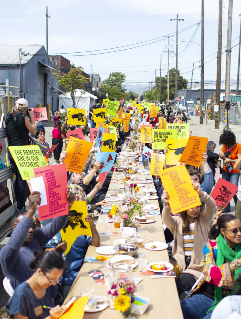People sitting at a very long table set for a meal; people are holding signs in many colors.