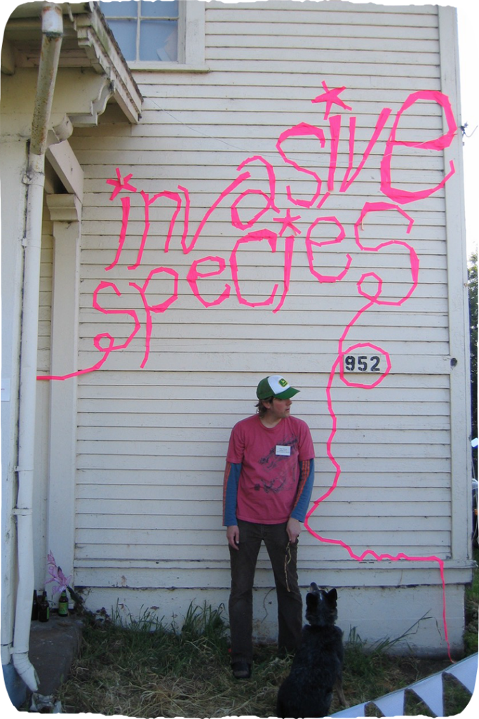 A person standing in front of a white building, the building scrawled with pink text that reads "invasive species"