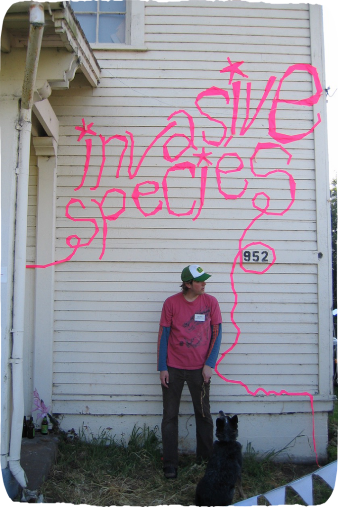 A person standing in front of a white building, the building scrawled with pink text that reads "invasive species"