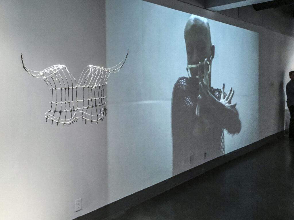 Photograph of a video projection featuring a person in a mask; a sculpture resembling a tunic made of wire hangs in the foreground