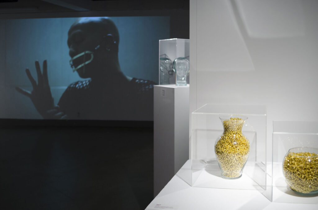 A photo of cars of yellow objects in the foreground, a video projection in the background with a masked figure