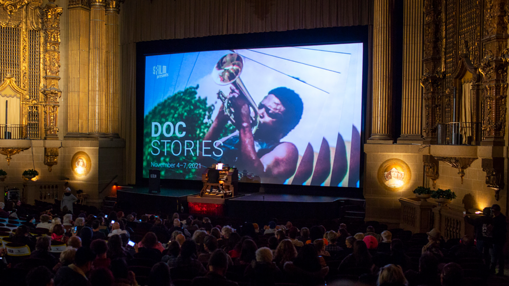 A theater with audience and a large screen with a repeating image of a person playing ahorn