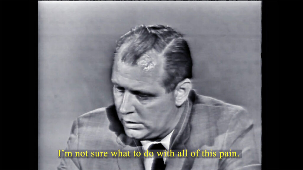 Black and white video still of a man in a suit with overlaid text that says 