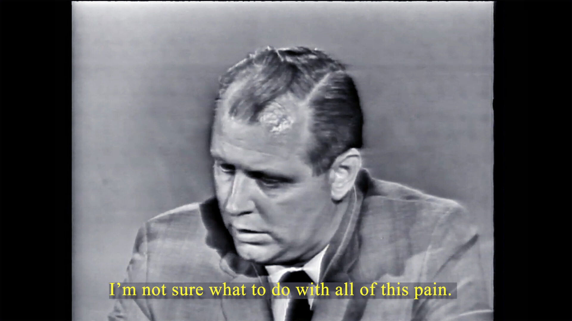 Black and white video still of a man in a suit with overlaid text that says
