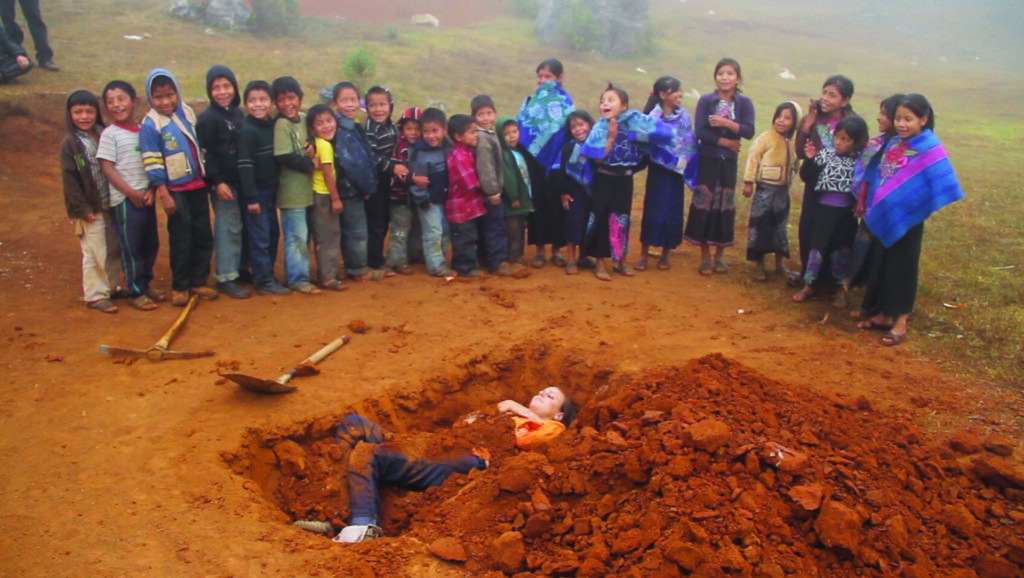 A group of people stand around a hole in the ground in which another person lies, partially covered with dirt.