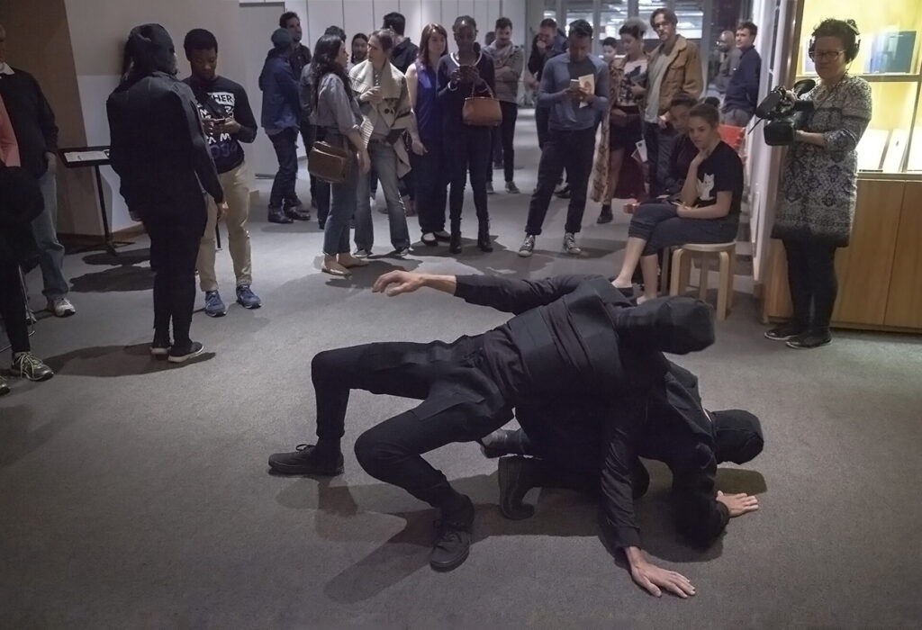 An audience watching two figures dressed in black velcro suits and masks colliding with each other.