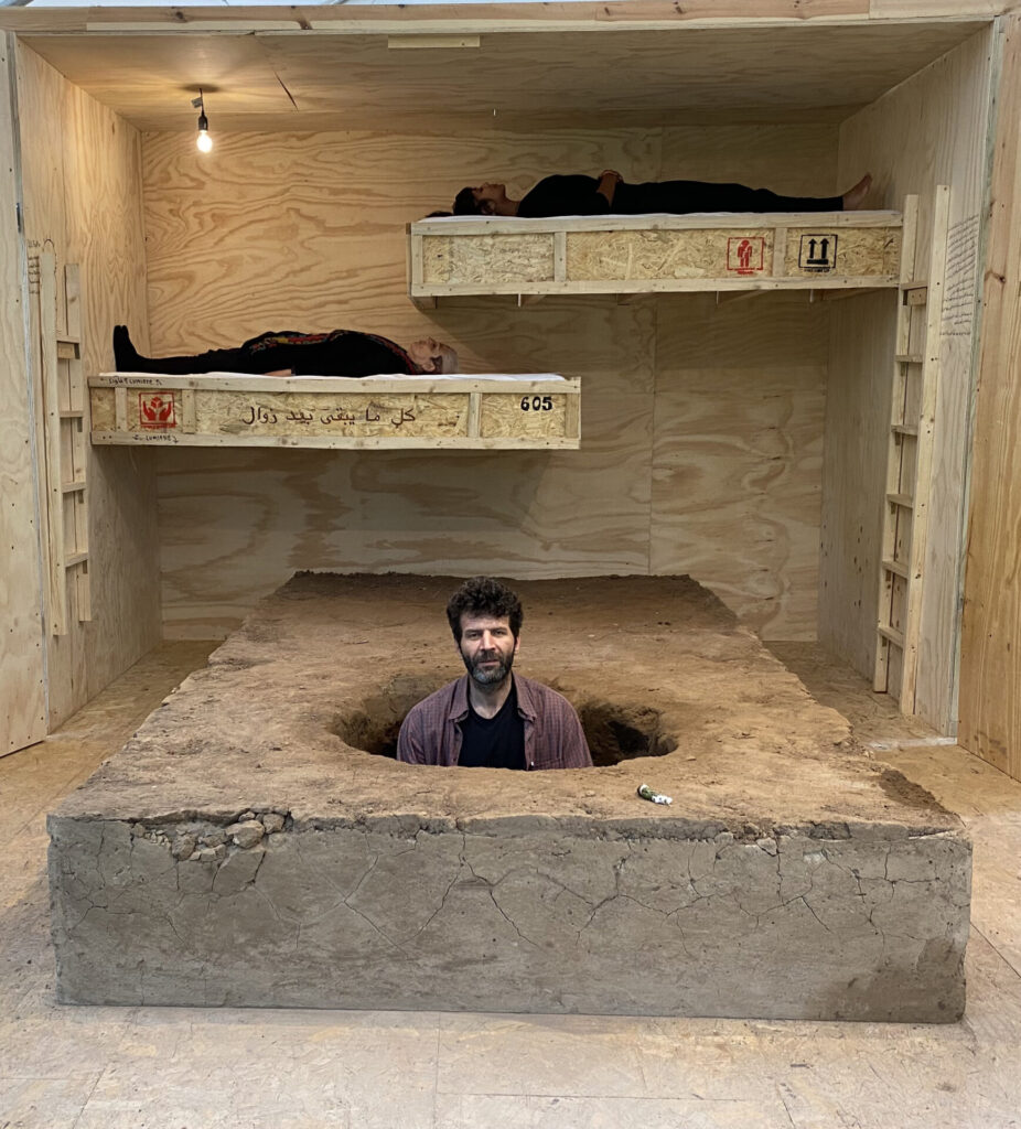 A room that appears to be made of dirt, with bunks on a far wall, and a person emerging from a round hole in the ground