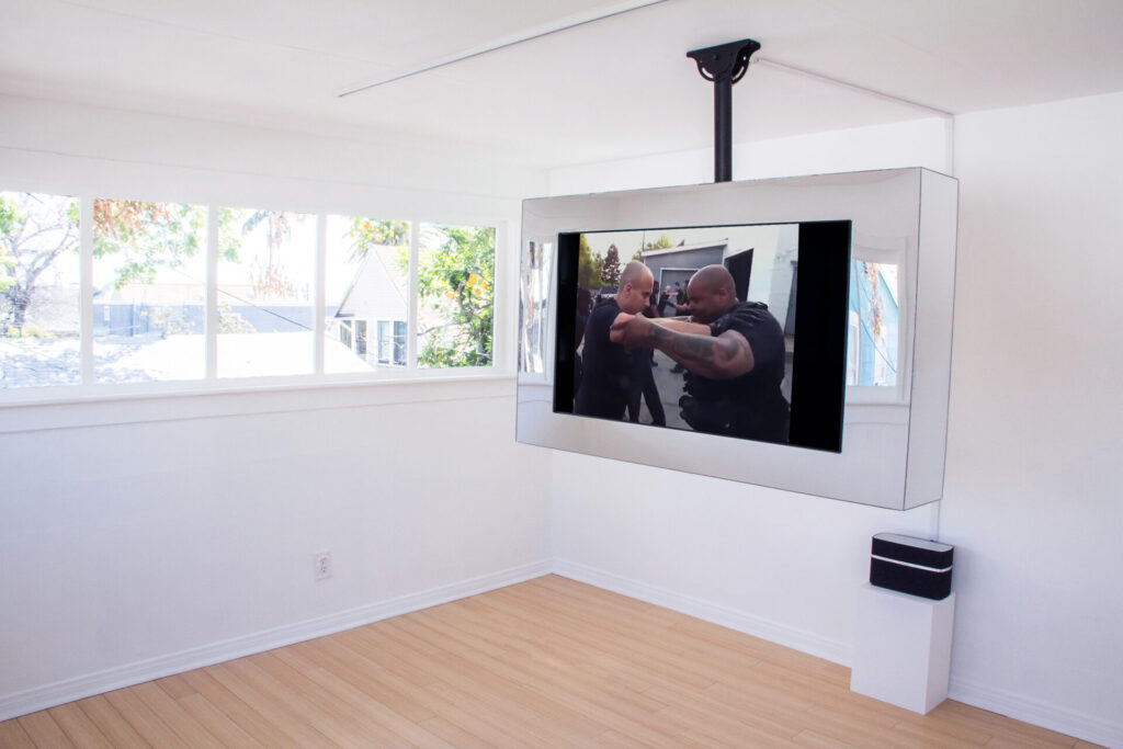 A video installed in a mirrored box suspended from the ceiling. The video depicts two men, one black and one white, arms interlocked as they engage in self-defense training.