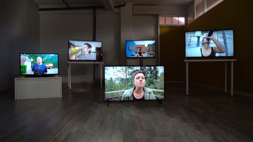 Installation view of five monitors of varying sizes displayed at different heights. The videos depict different people drinking water, or with water spilling down their chin or shirt.