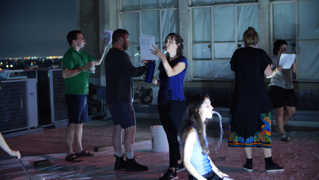 A group of people on a rooftop terrace engaged in various stages of drinking and spitting up water.