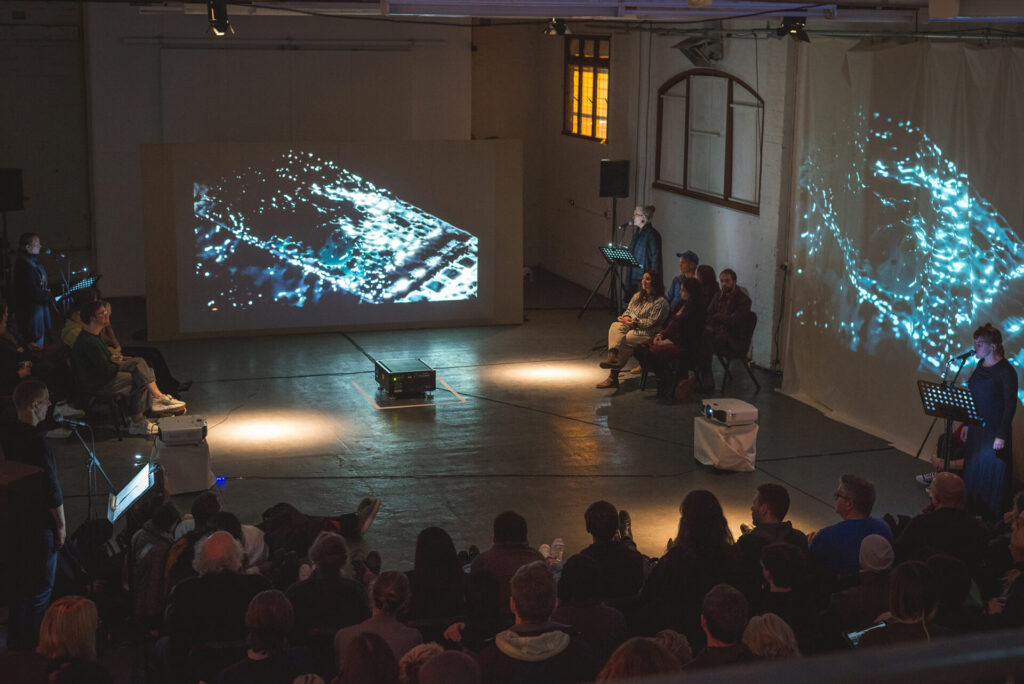 A group of people sitting on a stage with projections and an audience.