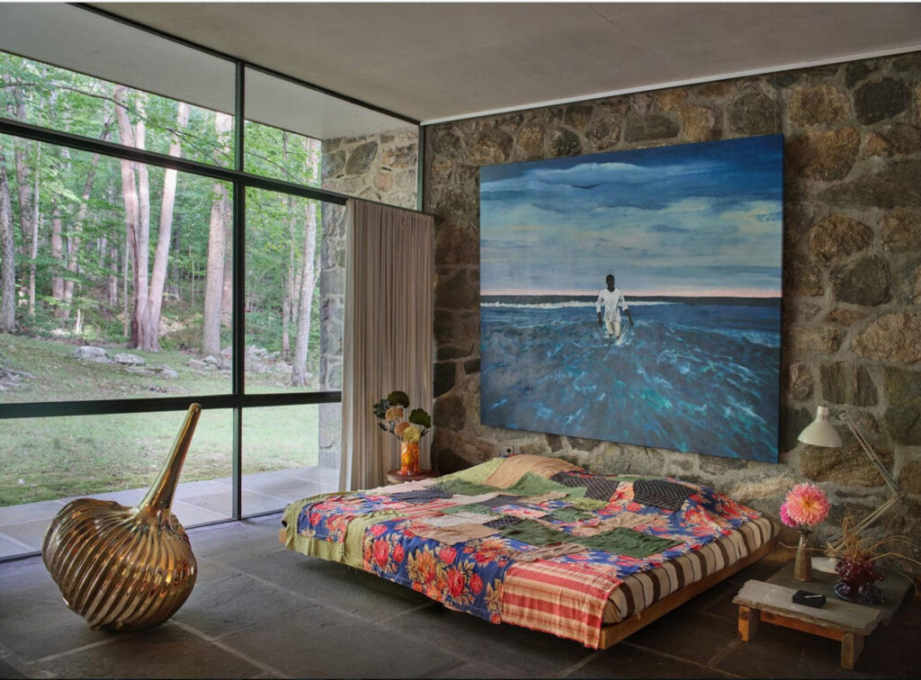 A photo of a room with a metal sculpture, a painting of a perso wading in ocean waves, and a large bed or couch with a red tiled quilt.