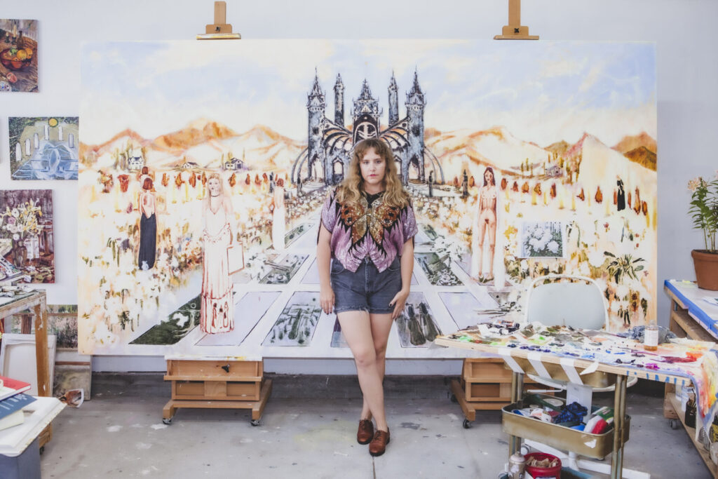 A woman, Shanna Waddell, wearing denim shorts and a sequined shirt with a butterfly, stands in front of a large painting set up across two easels. The painting depicts women both clothed and naked standing in a garden; a castle-like structure appears in the center, with mountains stretching behind it.