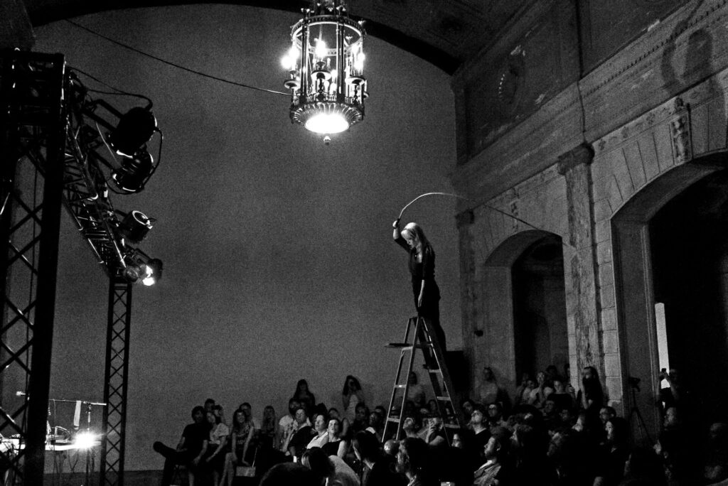 Black and white image of a woman at the top of ladder wielding a whip while a crowd looks on from below.