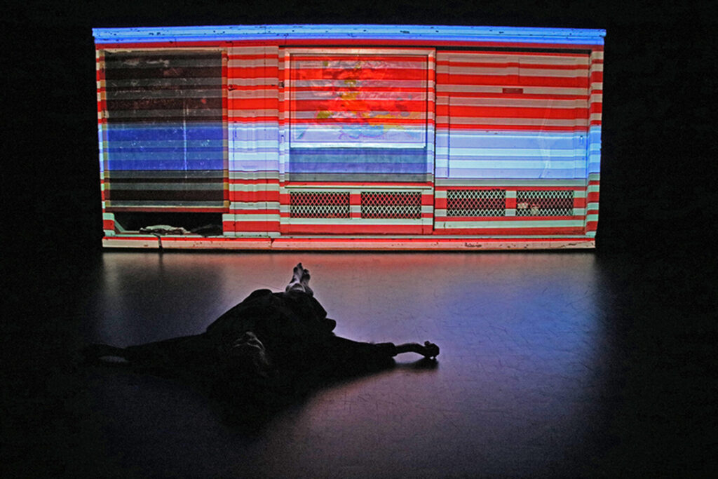 A figure lying on the floor in front of a large projection in blue and red