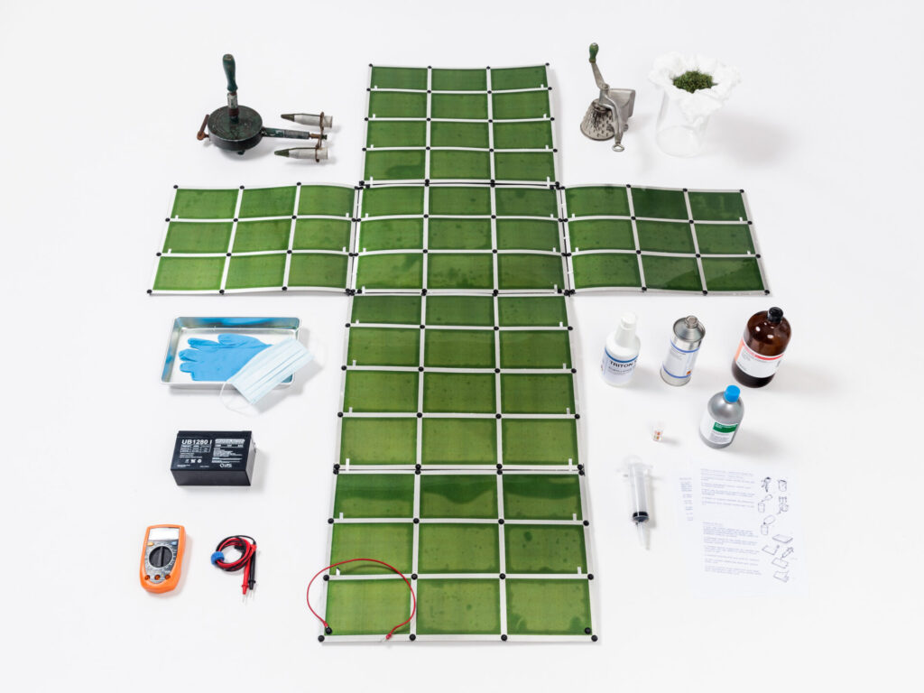 Gridded green paper arranged in the form of a cross, with tools around it including nitrile gloves, chemicals, voltmeter, and others.