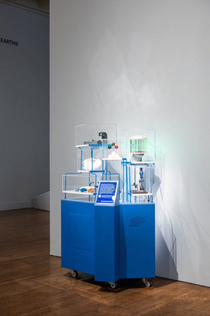 A blue pedestal on casters with various objects in plexiglass containers.