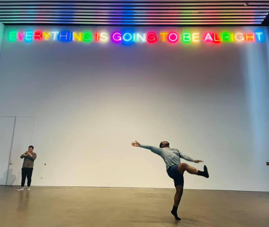 Dominic Moore-Dunson (a black man with short black hair) dancing and Tommy Lehman (a white man with short brown hair) playing trumpet in front of a white gallery wall with Martin Creed’s Work No. 203 art installment that reads in neon letters, “EVERYTHING IS GOING TO BE ALRIGHT.’