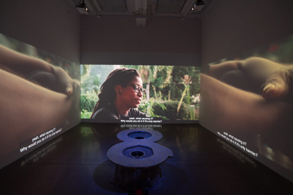A room with video projections on three walls depicting a person and folded arms, with a metal sculpture in the center