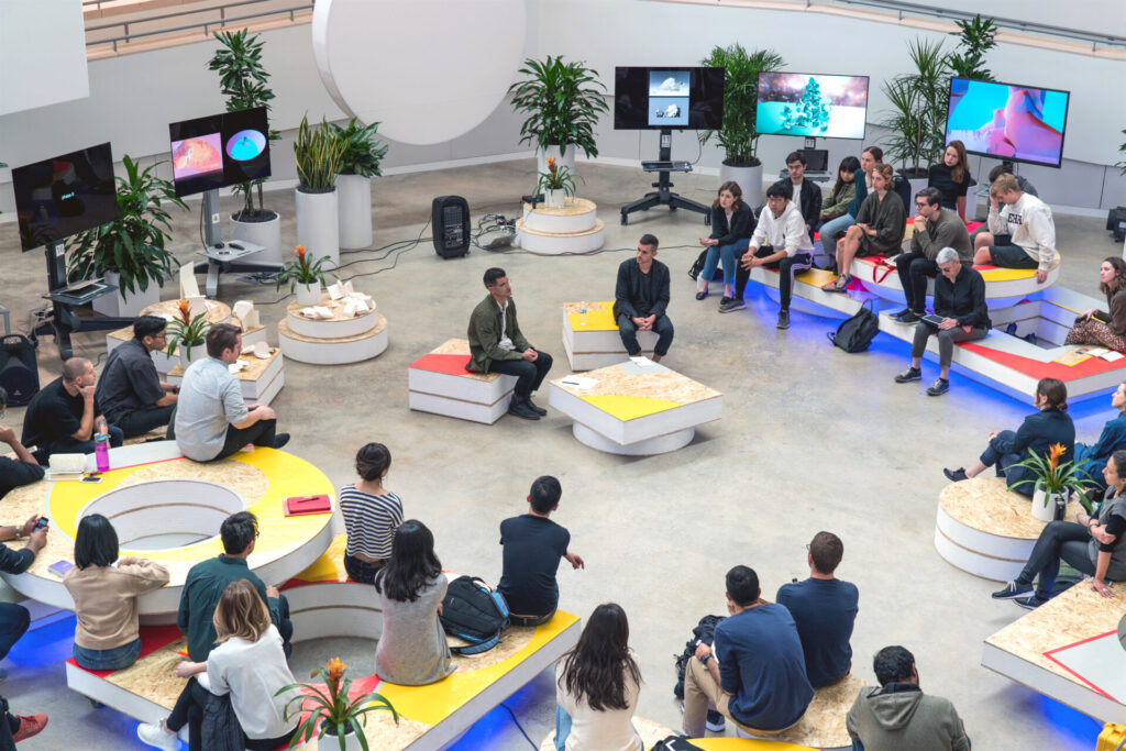 People sitting in a room furnished with multi-colored round and square platforms, with potted plants and tv monitors surounding the room.