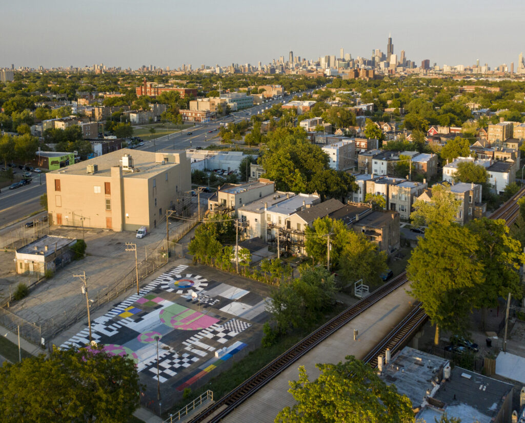 An aerial photograph of a city, with a basketball court in the foreground painted with patterns of black, white, green, and pink