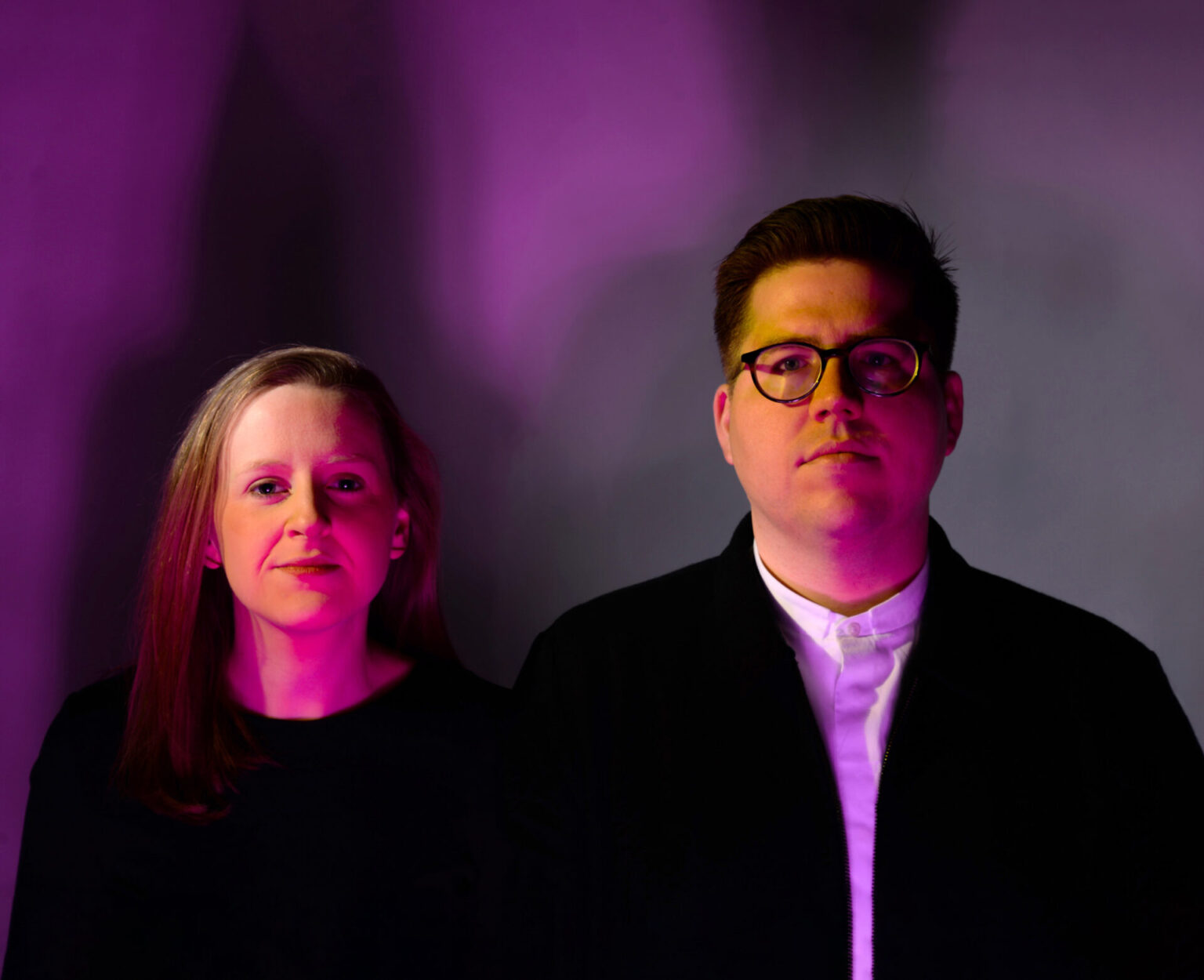 Portrait of Ashley Bigham and Erik Herrmann, both wearing black, photographed in pink lighting
