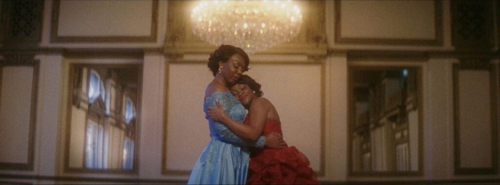 Two Black women in ball gowns hugging beneath a chandelier