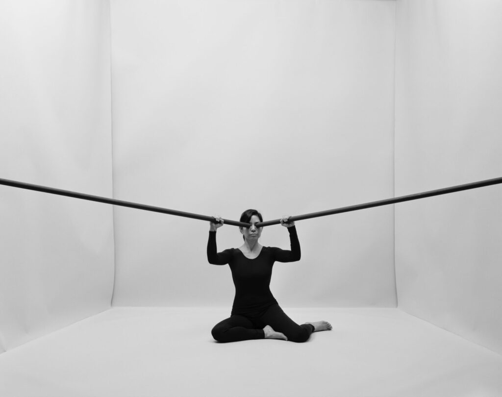 Image of a person dressed in black sitting on the floor in a white room, black rods pressed against their eyes, protruding out of the image frame