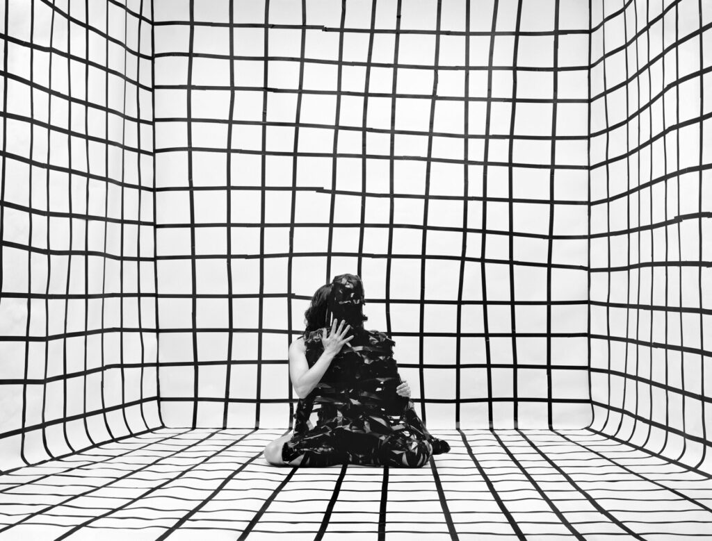 Photo of a figure seated on the floor of a room covered win a black and white grid. The figure holds another figure formed out of black facets.