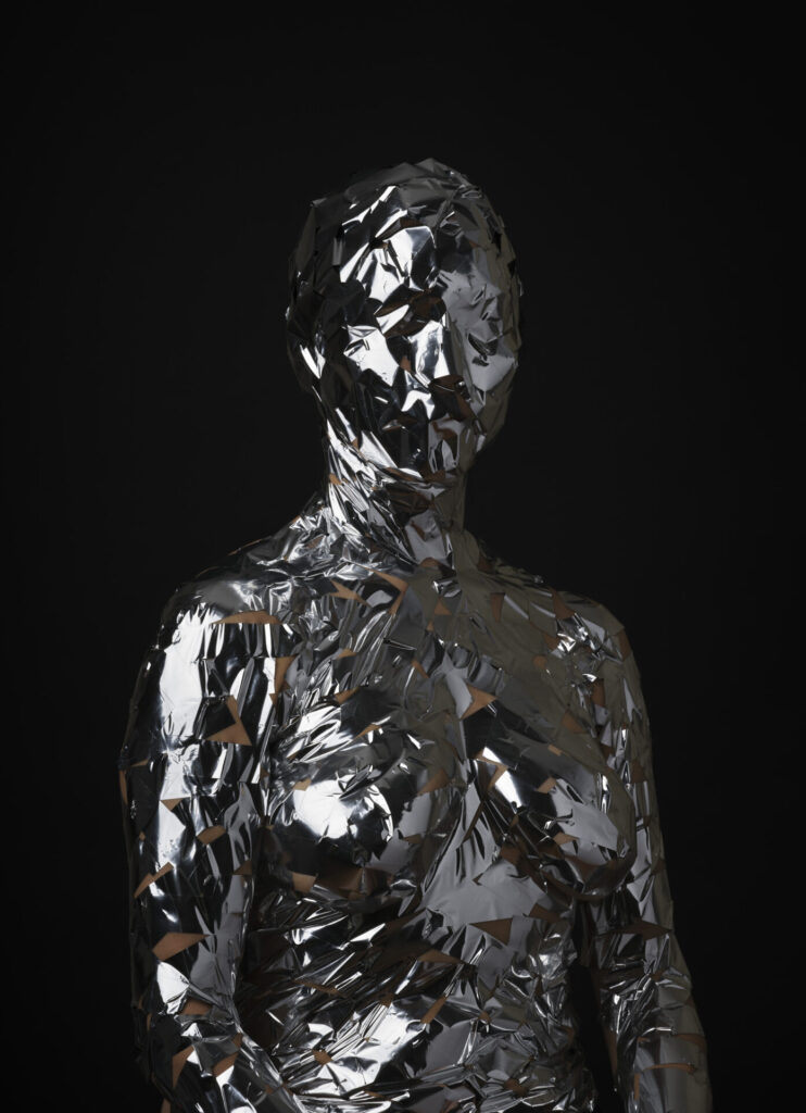 A photograph of a figure seen from the waist up, unclothed, wrapped in silver mylar tape, against a black background.