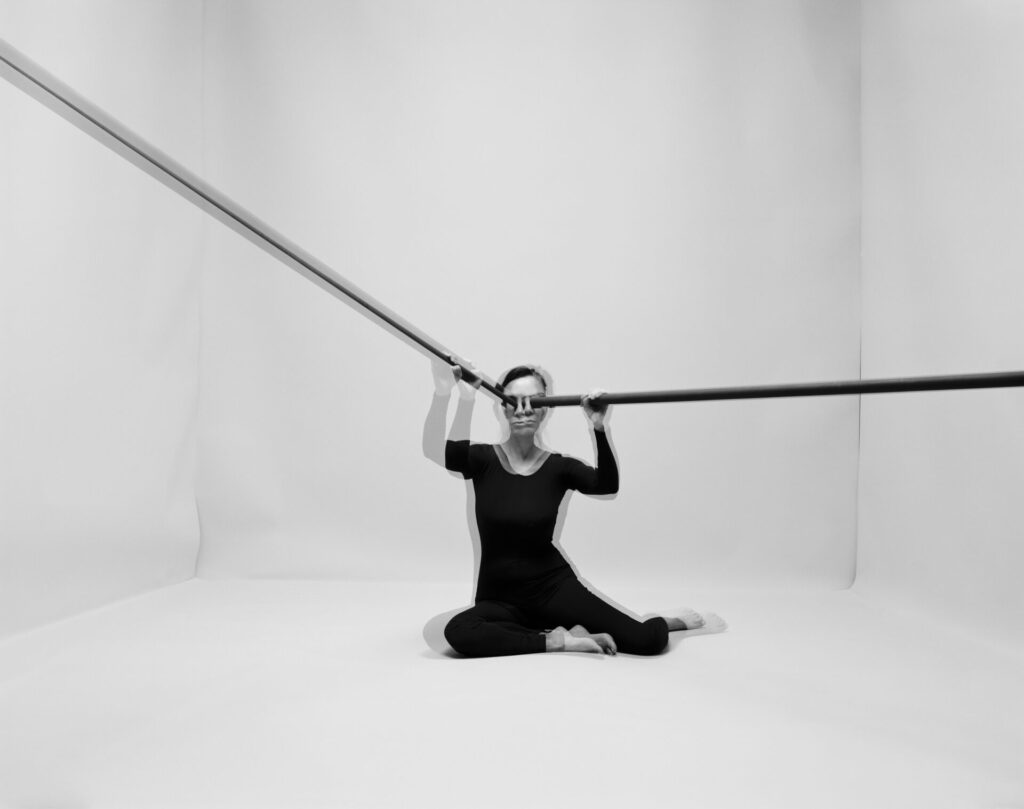 Image of a person dressed in black sitting on the floor in a white room, black rods pressed against their eyes, protruding out of the image frame. Th eimage is blurred or double-exposed, evoking a trembling movement.