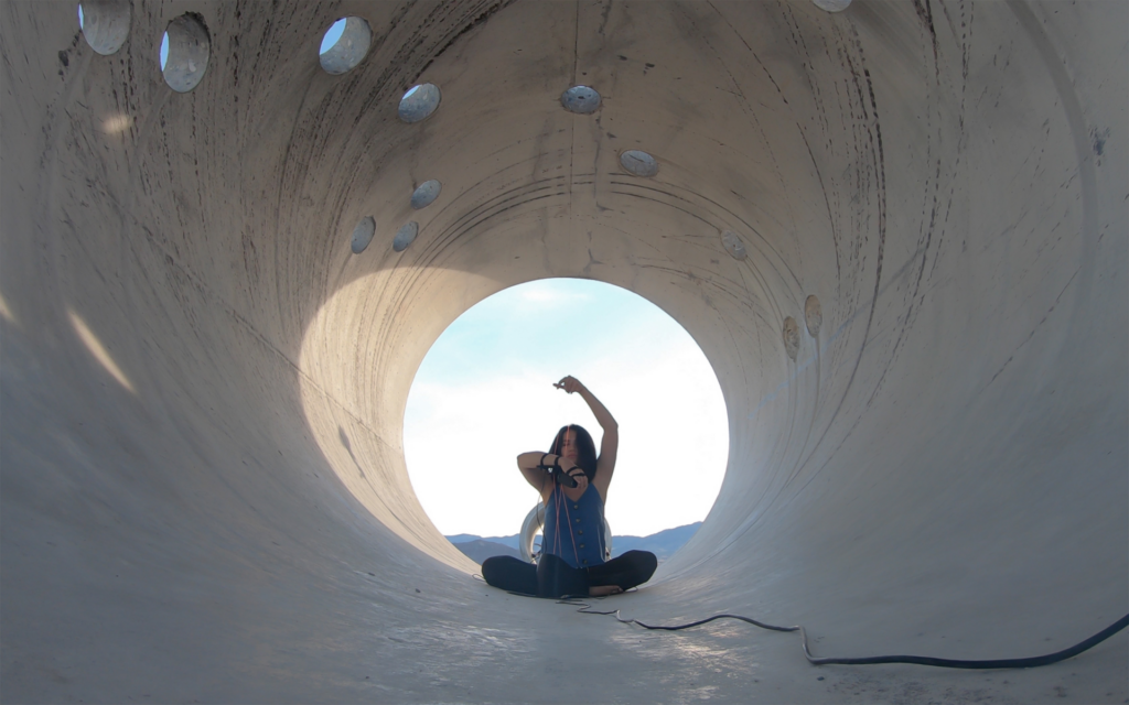 A photo of a person sitting in a concrete tube perforated by holes, arms raised, a wire extending toward the camera
