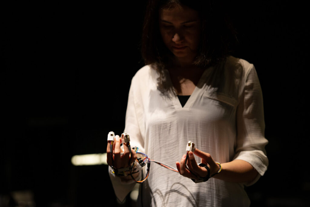 A person standing partially in shadows, hands held in front of them with multiple wires attached to their fingertips.