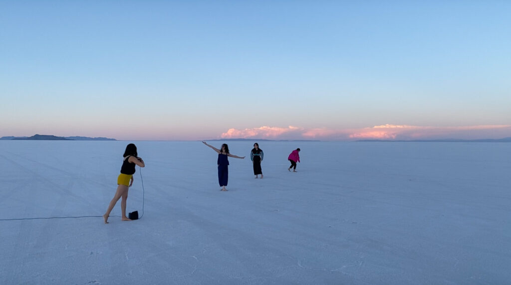 A line of people standing on pale flat ground, low clouds and blue sky in the far distance