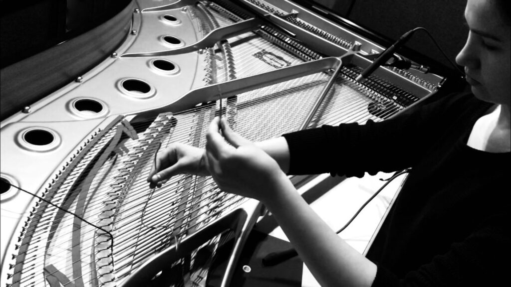 A black and white photo of hands manipulating the interior strings of a piano.