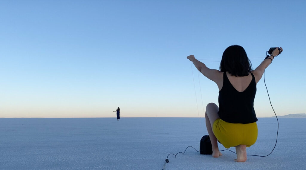 A photo with a person crouched in the foreground, wire extending and draping to the ground from extended arms; a person stands in the distance across a vast white plane.