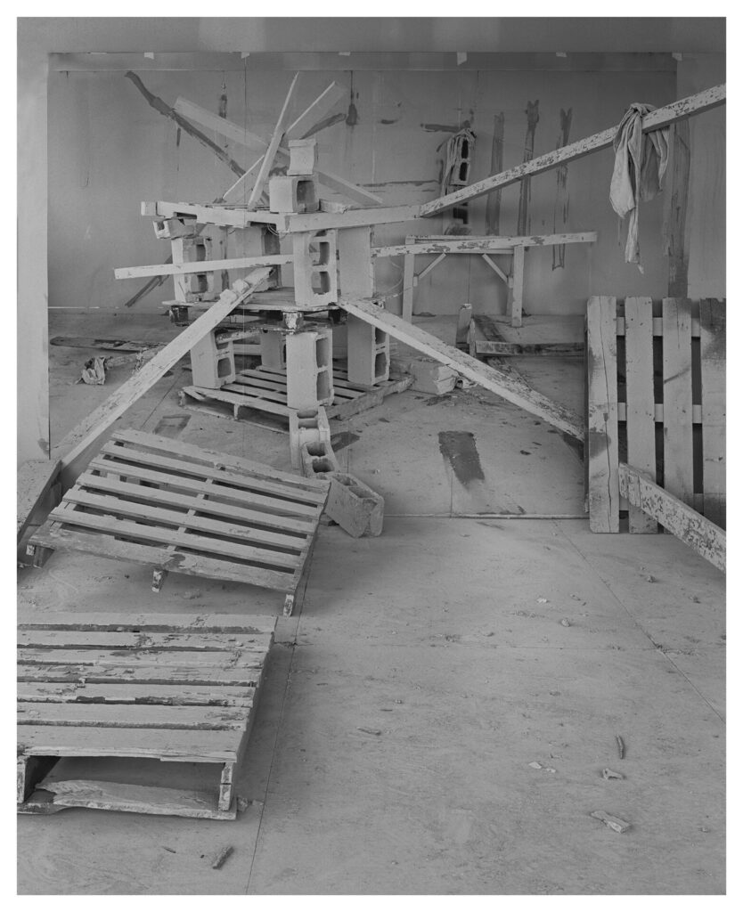 A black and white image of wooden pallets, cinder blocks, beams, and a rag arranged sculpturally in a raw space covered in dust.