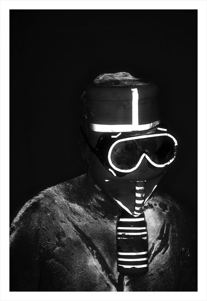 Black and white image of a person covered in a gas mask-like face covering and helmet with reflective tape.