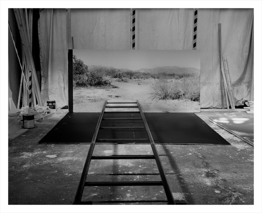 Black and white photo showing a large desert landscape photo standing upright on the ground, with a ladder laying perpendicular to it. Both are surrounded by paint cans and tarps.